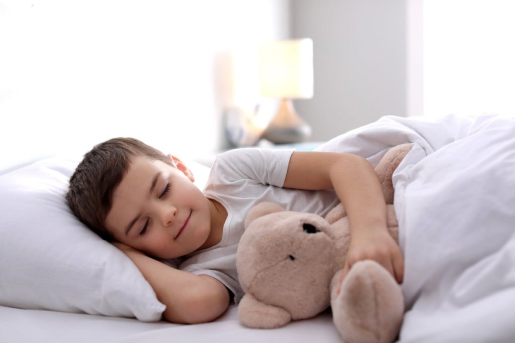 un petit garçon dort dans son lit avec un ours en peluche.