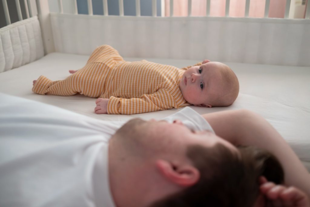 un papa endormir sur un lit dort aux côtés d'un bébé allongé dans son lit en cododo. Le bébé a les yeux ouverts et porte un pyjama à carreaux jaunes et blanc.