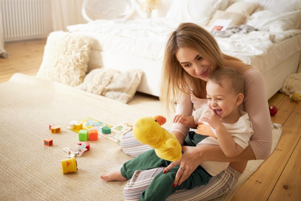 un jeune fille joue avec un enfant dans ses bras. il rit aux éclats devant une marionnette canard qu'elle tient devant lui. ils sont assis sur un tapis dans une chambre. divers jouets sont disposés sur le tapis.