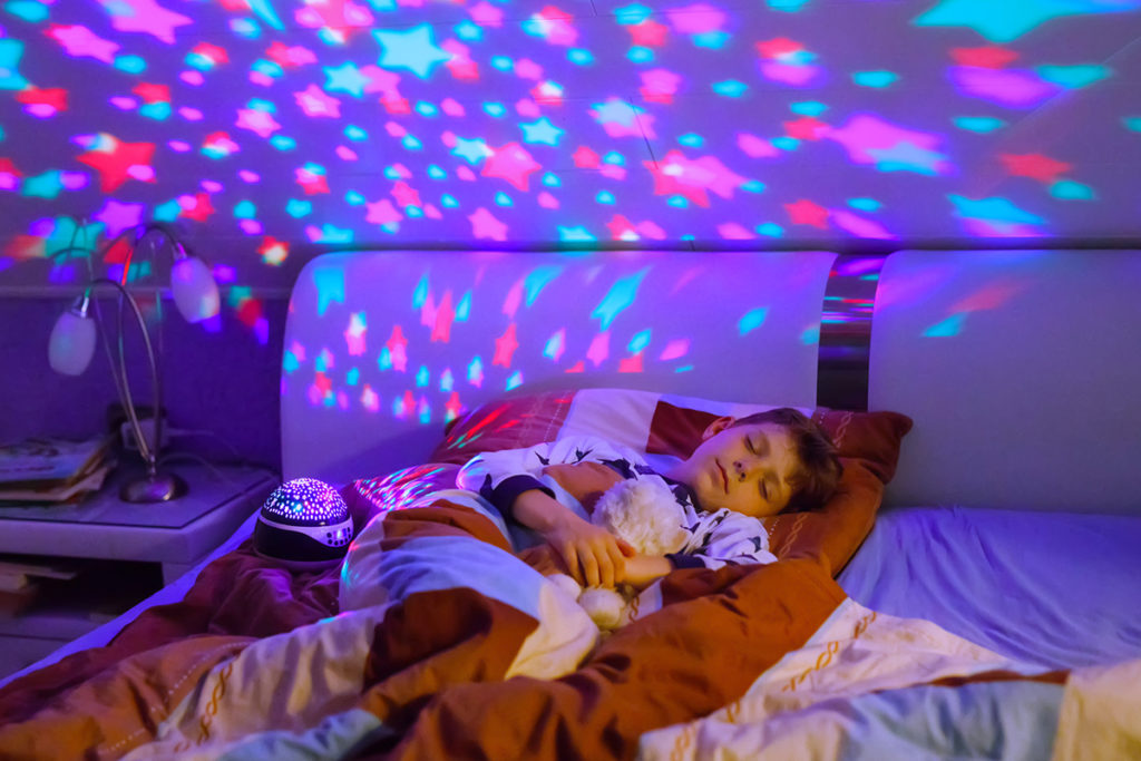 un enfant dort dans un lit avec son doudou dans les bras. une veilleuse allumée projette des étoiles roses et bleues sur le mur et le plafond.