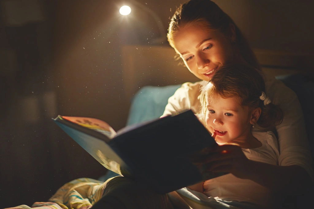 une jeune fille est assise dans les bras de sa maman qui lui lit un livre à la lumière d'une lampe de chevet.