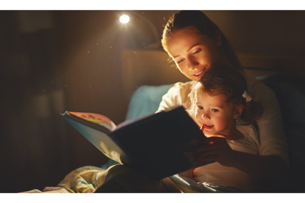 assises dans un lit, une maman et sa petite fille lisent un livre ensemble à la lumière d'une lampe de chevet.