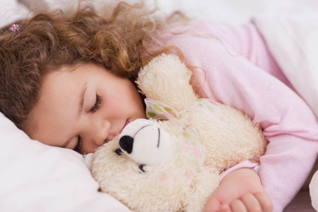 dans un lit, une petite fille dort avec son ours en peluche dans ses bras. elle porte un haut rose.