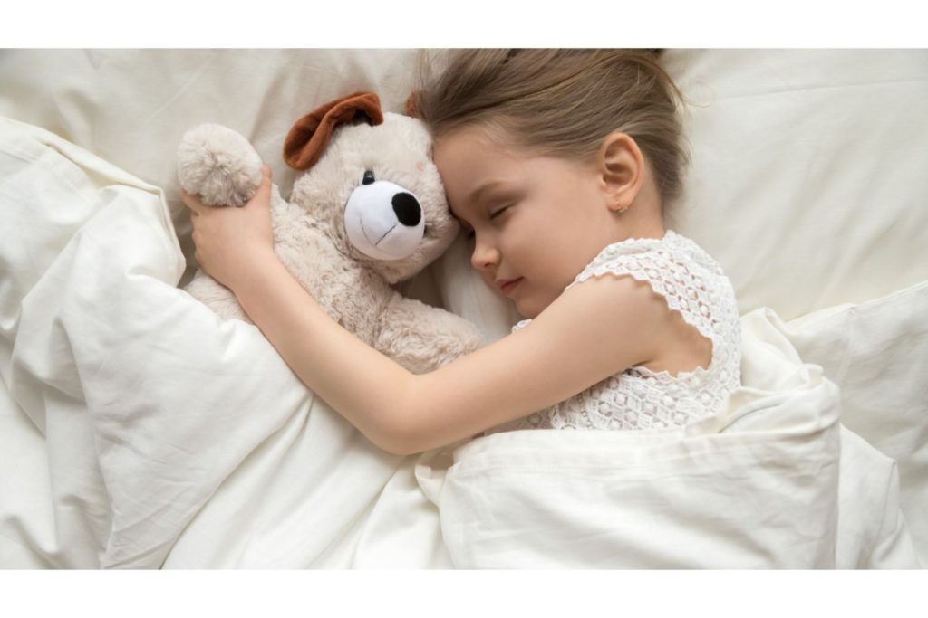 dans un lit blanc, une petite fille dort avec son ours en peluche contre elle.