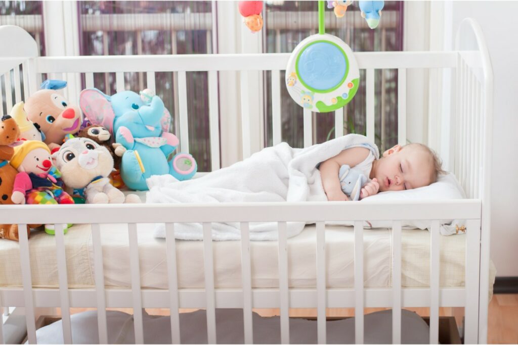 un bébé dort dans un lit à barreaux blanc. une veilleuse est suspendue au-dessus de lui. des peluches sont dans le lit au fond du lit.