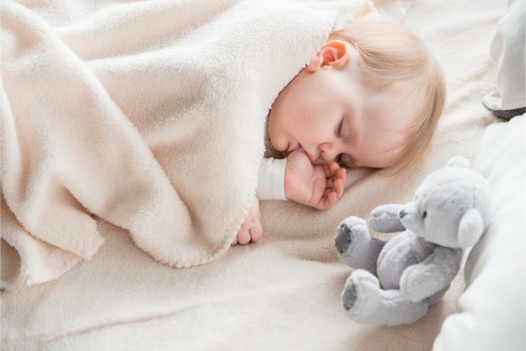 un bébé sous une couverture suce son pouce en dormant. une peluche nounours gris est posé à côté de lui.