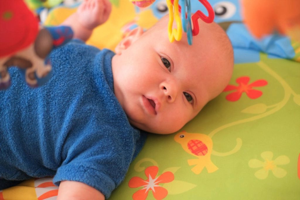 un bébé est allongé sur un tapis de jeu. il porte un habit bleu et regarde le photographe.
