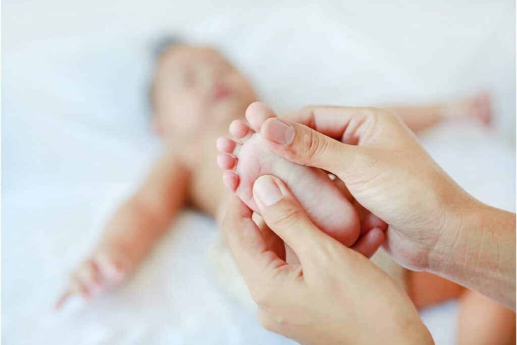 focus sur un pied de bébé avec 2 mains qui massent le pied du bébé. en fond flouté, on voit le bébé allongé les bras écartés.