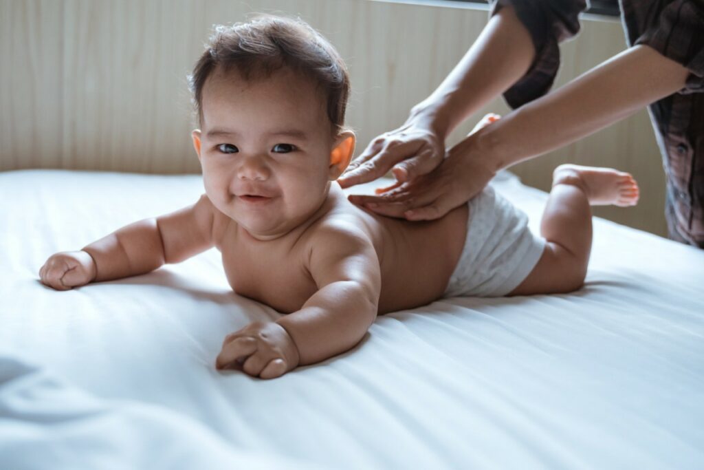 un bébé sur le ventre sur un lit, regarde le photographe pendant que des mains lui massent le dos.