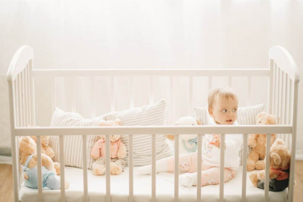 un bébé est assis dans un lit à barreaux blanc. 3 coussins à rayures sont disposé dans le lit ainsi que 6 ours en peluche.