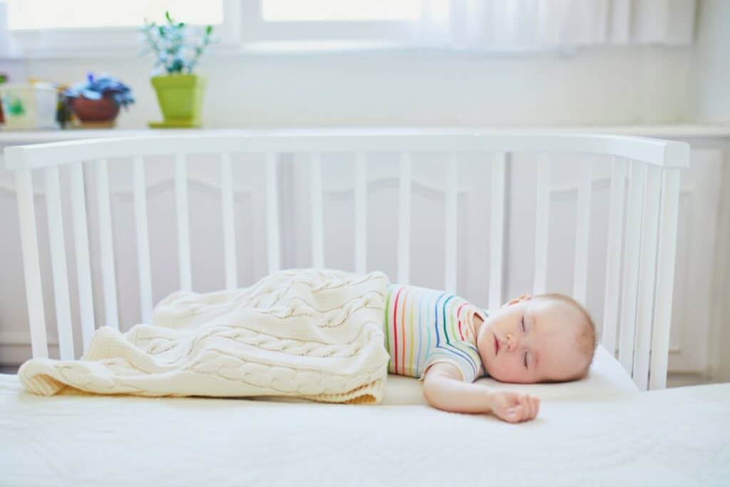 un bébé est endormi dans un lit cododo blanc collé au lit des parents. il porte un body manches courtes à rayures. il a sur lui un couverture beige.