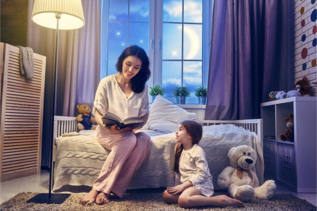 dans une chambre d'enfant au clair de lune, une jeune fille est assise au sol et regarde sa mère assise sur le lit qui souriant avec un livre ouvert.