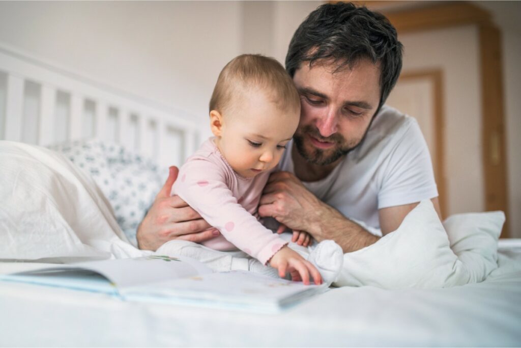 sur un lit, un papa et son bébé sont posés et lisent ensemble un livre. la petite attrape la page du livre pour la tourner. elle porte un body rose et son papa un tee-shirt blanc.