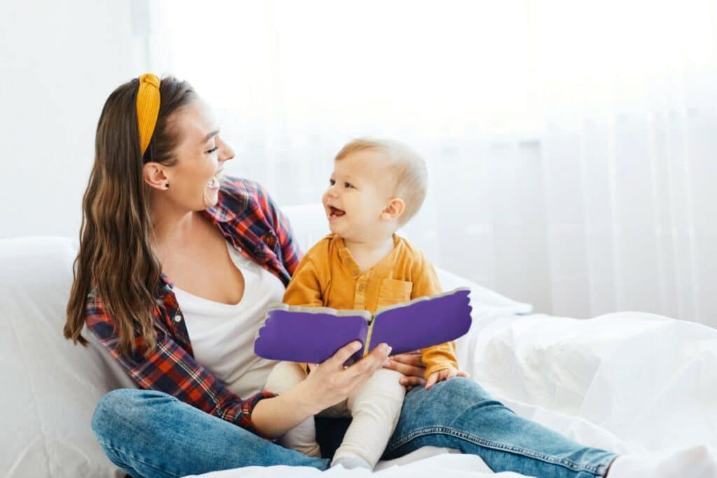 sur un canapé blanc, une mère est assise avec son enfant sur les jambes. elle tient un livre ouvert devant son enfant. ils se regardent en souriant l'un l'autre.