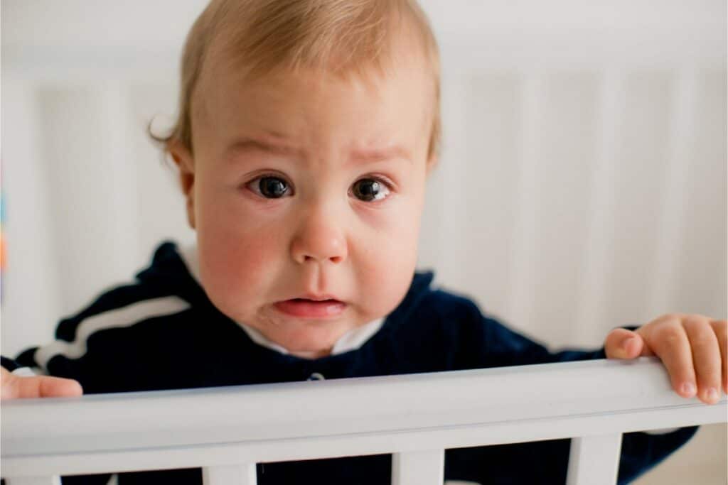 plan rapproché sur un bébé qui a les larmes aux yeux et tient son lit à barreaux avec ses deux mains.