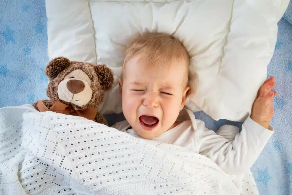 dans son lit, la tête sur un oreiller plat blanc et sous une couverture blanche, un bébé pleure. il a son nounours à côté de lui.
