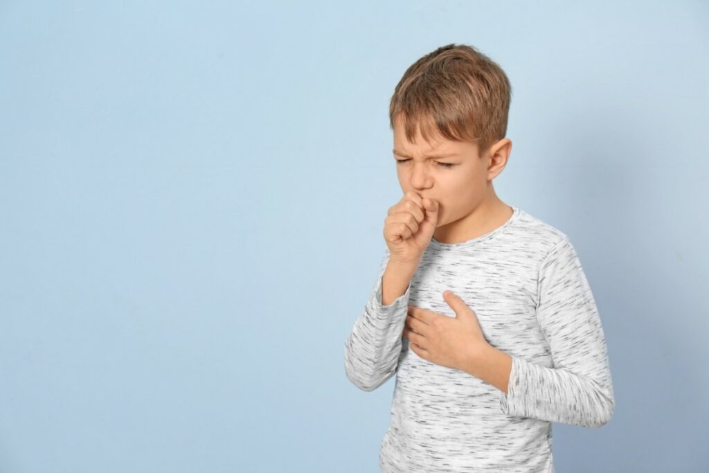devant un fond bleu ciel, un enfant debout tousse avec une main devant la bouche et l'autre sur la poitrine.