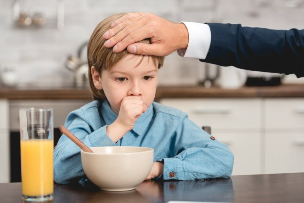 un enfant tousse à table devant son bol et son verre de jus d'orange. il met sa main devant la bouche. une main d'homme en costume lui touche la tête.