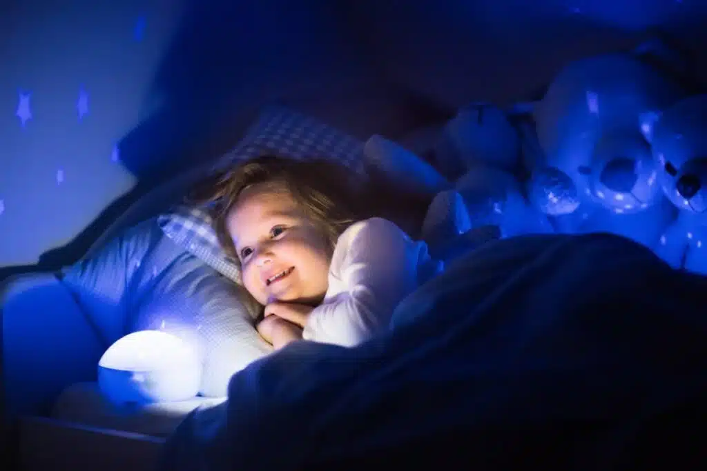 une petite fille est couchée dans son lit. sa veilleuse projette des étoiles partout dans sa chambre dans la nuit. elle sourit couchée dans son lit.