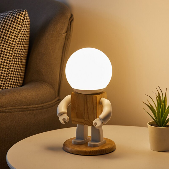 Veilleuse lampa allumée en forme de petit robot en bois et métal dont la tête est la lampe allumée , elle est posée sur une petite table près d'un fauteuil , à côté d'elle se trouve une mini plante verte dans un tout petit pot