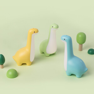 Trois veilleuses en forme de dinosaure, une jaune, une bleue et une verte posées sur un fond vert avce des petits arbes en bois.