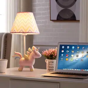 sur un bureau près d'un ordinateur portable ouvert, et d'un petit pot de fleurs, une lampe dont la base est une licorne rose avec la queue couleur arc en ciel